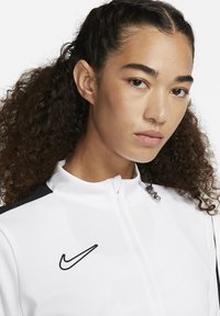 Valkoinen vetoketjullinen takki mustilla yksityiskohdilla ja musta Nike-logo rinnassa, jossa on korkealle ulottuva kaulus ja sileä pinta.