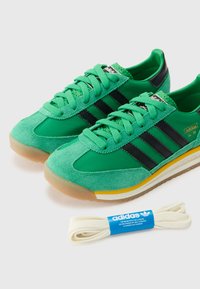 Tênis verdes de camurça e têxtil com riscas pretas, cordões texturizados, sola de borracha e detalhes amarelos. Inclui um cordão branco adicional.