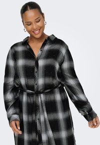 Smiling woman with hoop earrings wearing a black and gray plaid belted shirt dress, posing with eyes closed against a plain background.