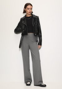 Černá kožená zkrácená bunda přes černý top, šedé střižené široké kalhoty a černé boty s bílými akcenty, postavená před neutrálním pozadím.