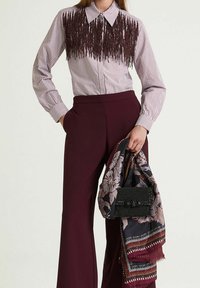 Camicia a righe con bottoni e dettaglio in frange di colore rosso scuro, abbinata a pantaloni bordeaux a gamba larga e una sciarpa fantasia, tenuta nella mano sinistra.