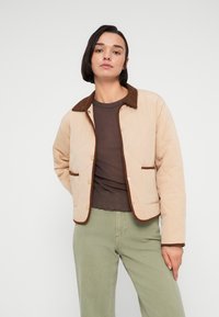 Μπεζ μπουφάν με επένδυση, καφέ γιακά και διακοσμητική τρέσα; διαθέτει κουμπιά τύπου σναπ και πλαϊνές τσέπες; φορεμένο πάνω από ένα καφέ τοπ και πράσινο παντελόνι.