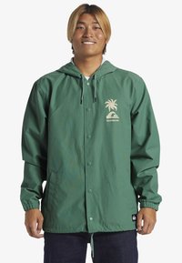 Grøn hættejakke lavet af let materiale, med et palmetræ-logo og et Quiksilver-mærke. Elastiske manchetter og en knaplukning.