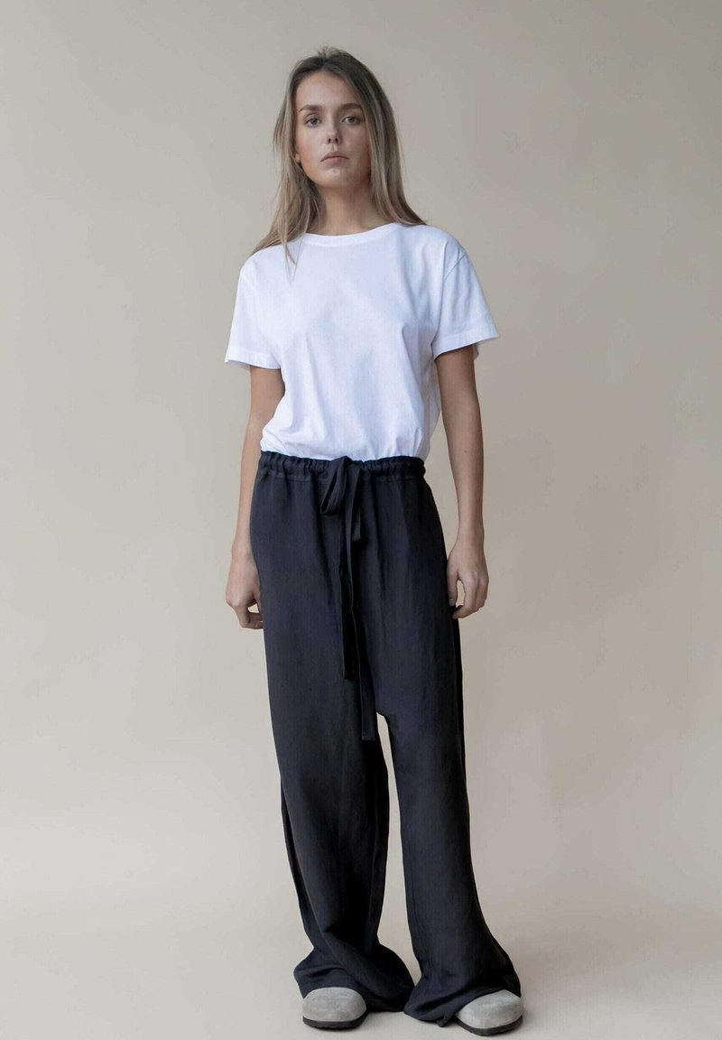 Witte katoenen t-shirt gecombineerd met donker, losvallende broek met elastische taille. Zachte textuur en subtiele glans. Model draagt grijze schoenen.