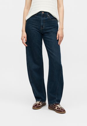 Άτομο που φοράει σκούρο μπλε τζιν με ψηλή μέση και ίσια μπατζάκια, λευκή αμάνικη μπλούζα με ρίγες και καφέ αθλητικά παπούτσια με λευκά κορδόνια σε λευκό φόντο.