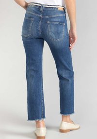 Jeans en denim de bleu moyen, présentant une coupe droite, des ourlets effilochés et deux poches arrière. Portés avec des chaussures de couleur claire.