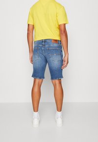 Ber shorts in denim azzurro chiaro con orlo sfilacciato. Presentano due tasche posteriori e un'etichetta in pelle marrone sulla vita. Indossati con sneakers bianche.