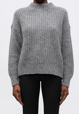 Personne portant un pull en maille texturée gris à manches longues et un pantalon noir, debout devant un fond blanc uni.
