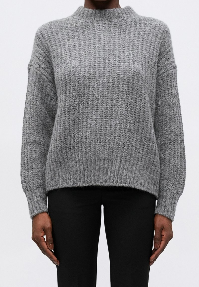 Personne portant un pull en maille texturée gris à manches longues et un pantalon noir, debout devant un fond blanc uni.
