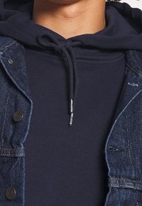 Felpa con cappuccio blu navy con coulisse, indossata sotto una giacca di jeans. La giacca presenta bottoni in metallo e un tessuto con texture.