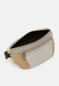 Heuptasje in beige en tan, met een gaas voorvak, ritsluiting en stoffen band. Gladde textuur met subtiele patronen.