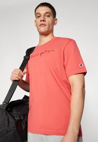 T-shirt en coton corail avec logo "Champion" brodé, col rond, manches courtes et design ajusté ; associé à un sac de sport noir.