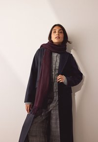 Tumšs mētelis pār pelēku džemperi, kombinēts ar vīnsarkanu šalli un rūtainām biksēm. Mētelim ir pielāgota piegrieztne, bet šallei ir malās pušķi.