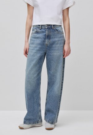 Person trägt locker sitzende hellblaue Jeans mit weißem T-Shirt und weißen Turnschuhen, steht vor einem einfarbigen Hintergrund.