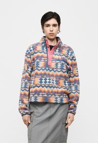 Άτομο που φοράει πολύχρωμο φλις πουλόβερ με ροζ κουμπωτή λωρίδα και γκρίζα φούστα καράβι, στέκεται μπροστά σε απλό φόντο.