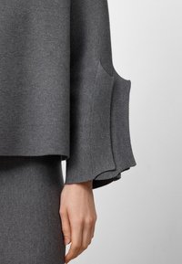 Top grigio in maglia con una vestibilità rilassata, caratterizzato da maniche larghe con bordi svasati e una texture liscia. Alcuni tessuti sono raccolti ai lati.