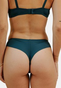 Bas de bikini teal avec une texture lisse, un design taille basse et une couverture minimale, présentant une finition sans couture et sans matériel visible.