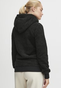 Sudadera negra y esponjosa con una superficie texturizada, que presenta una capucha con cordón y puños de canalé, combinada con pantalones de color claro.