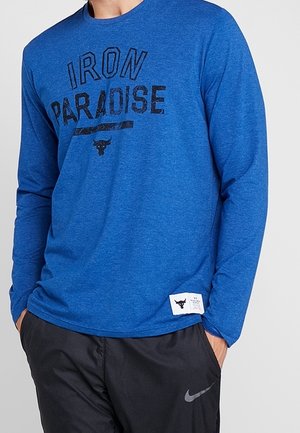 Mężczyzna ubrany w niebieską koszulę z długim rękawem z napisem „IRON PARADISE” oraz czarne spodnie sportowe z logo Nike, ręce w kieszeniach.