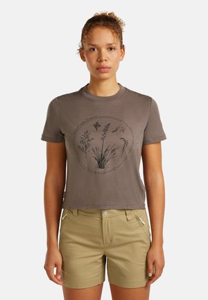 Kobieta o kręconych włosach w brązowej koszulce z krótkim rękawem z roślinnymi nadrukami i beżowych szortach, stojąca na jednolitym tle.