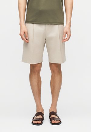 Άνδρας ντυμένος με μπεζ σορτς μήκους μέχρι το γόνατο, ελιάς πράσινη μπλούζα και σκούρα καφέ σανδάλια, που στέκεται μπροστά σε απλό φόντο.