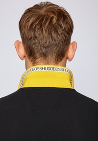 Homme aux cheveux bruns courts portant une chemise noire avec un col relevé jaune affichant le texte « HUGO BOSS » en répétition.
