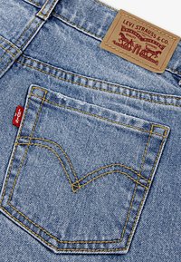 Jasnoniebieskie dżinsy z żółtym przeszyciem, posiadające tylnią kieszeń z charakterystycznym wzorem w kształcie litery V oraz brązową skórzaną naszywkę z logo.