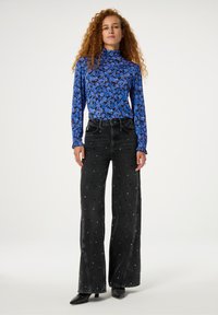 Langärmliges, blaues, floral gemustertes Rollkragenoberteil kombiniert mit hochgeschnittenen, schwarzen Jeans, verziert mit silbernen Verzierungen, und spitzen, schwarzen Schuhen.