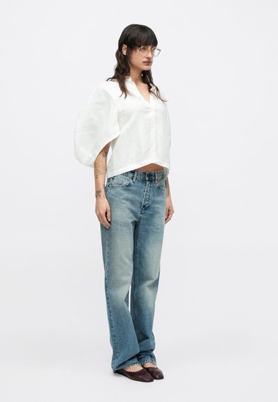 Λευκό, φαρδύ πουκάμισο με μεγάλα μανίκια, συνδυασμένο με ανοιχτό μπλε, ξεθωριασμένο τζιν με ευθεία γραμμή και μπορντό παπούτσια χωρίς κορδόνια.