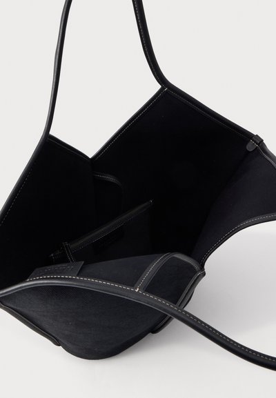Sac cabas en cuir noir avec longues anses, coutures blanches, ouverture sur le dessus laissant voir une poche intérieure zippée et une doublure unie.