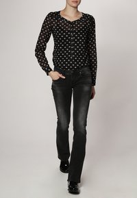 Blouse noire transparente à pois blancs, manches longues et détails à boutons, associée à un jean évasé foncé et des chaussures noires brillantes.