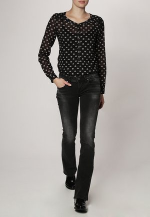 Zwarte doorschijnende blouse met witte stippen, lange mouwen en knoopdetails, gecombineerd met donkere uitlopende spijkerbroeken en glanzend zwarte schoenen.