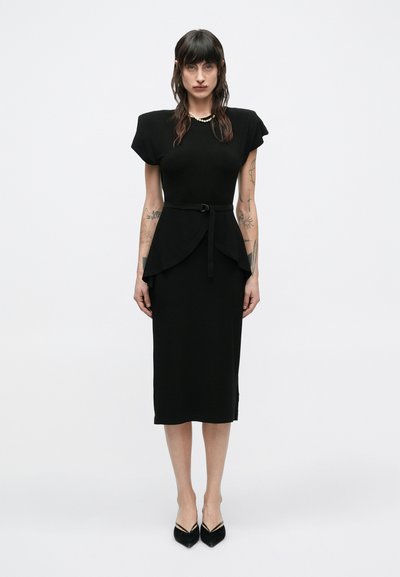 Femme aux cheveux foncés et tatouages, portant une robe midi noire avec des épaulettes, une ceinture à la taille, un collier de perles et des escarpins noirs à bout pointu, sur un fond blanc.