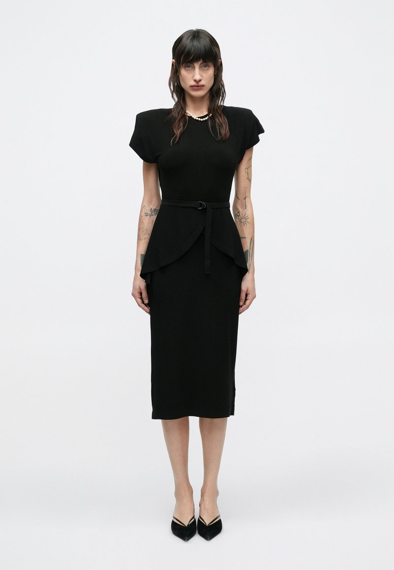 Kobieta o ciemnych włosach i tatuażach, ubrana w czarną sukienkę midi z poduszkami na ramionach, paskiem w talii, perłowym naszyjnikiem i czarnymi szpiczastymi szpilkami na białym tle.