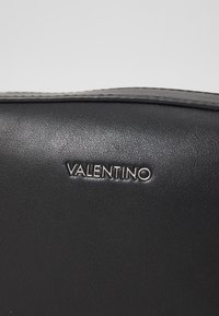 Sac en cuir noir avec une surface texturée, comportant le nom de la marque embossé "VALENTINO" en argent. Design simple et épuré.