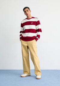 Sudadera a rayas rojas y blancas con cuello redondo y logo, combinada con pantalones de pierna ancha beige y zapatillas blancas con suelas de goma.