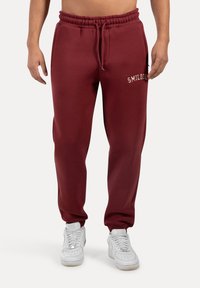 Bordeaux kleurige joggingbroek met elastische tailleband en trekkoord. Voorzien van een zijzak en logo detail op de linkerdij. Van katoenmix materiaal.