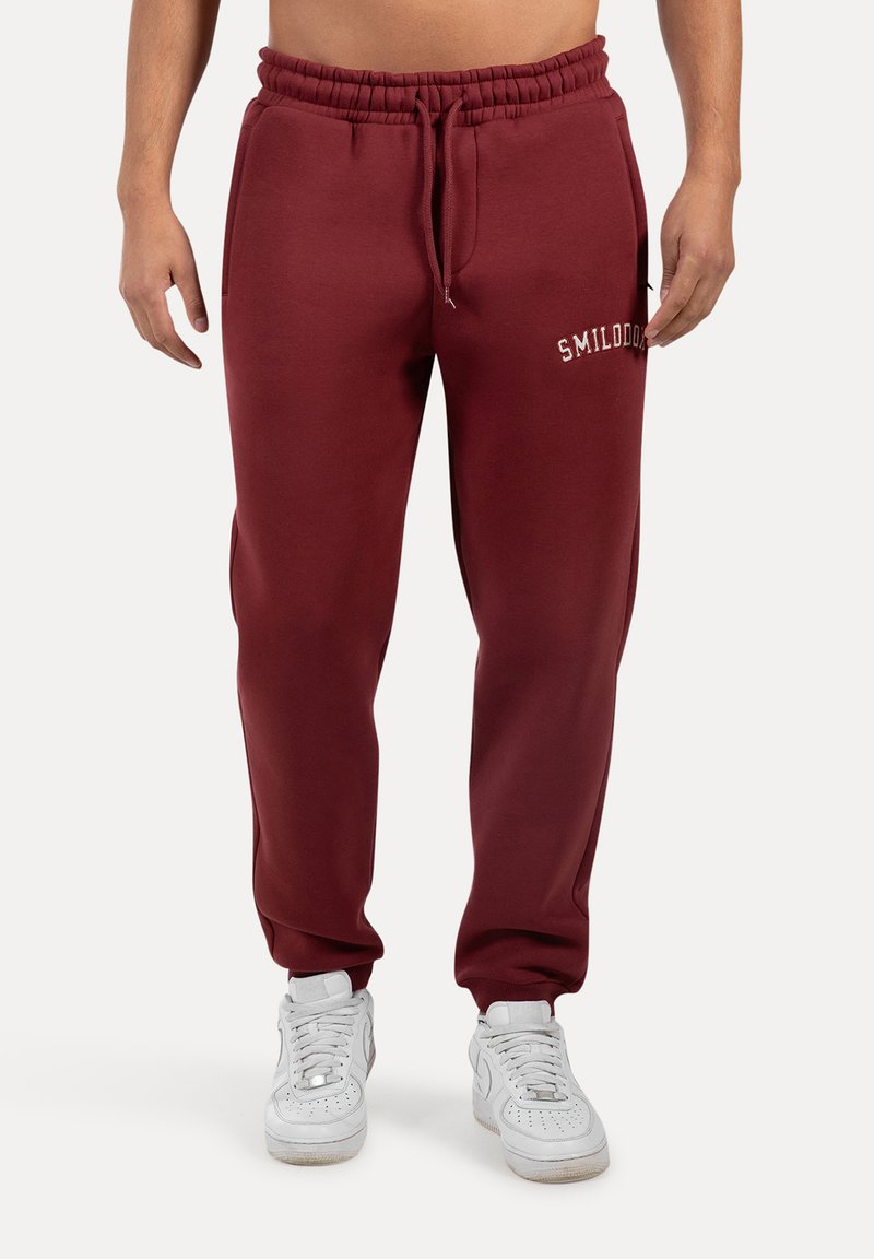 Bordeaux kleurige joggingbroek met elastische tailleband en trekkoord. Voorzien van een zijzak en logo detail op de linkerdij. Van katoenmix materiaal.