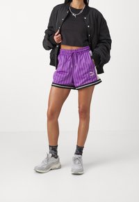 Personne portant une veste bomber noire, un crop top noir, un short en mesh violet Puma avec des bandes noires, des chaussettes noires et des baskets blanches, debout.