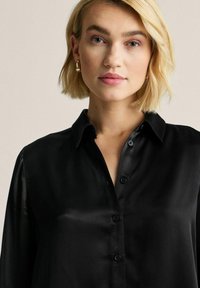 Sort satinbluse med klassisk krave, knaplukning foran og lange ærmer. Glat tekstur, med sorte knapper og minimale detaljer.