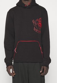 Schwarzer Hoodie mit roten Akzenten, der ein Grafik von einem Hunde Kopf auf der Brust zeigt. Er hat eine vordere Tasche und eine Kordelzugkapuze.