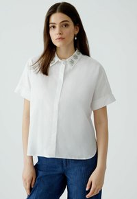Weißes, kurzärmliges Hemd mit Knopfleiste und spitzem Kragen, das dekorative Verzierungen aufweist. Glatte Textur und lockere Passform, kombiniert mit blauen Jeans.