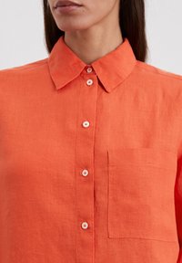 Camicia button-up in lino arancione con colletto, tasca frontale e orlo arrotondato. Presenta bottoni bianchi e un tessuto caratterizzato.