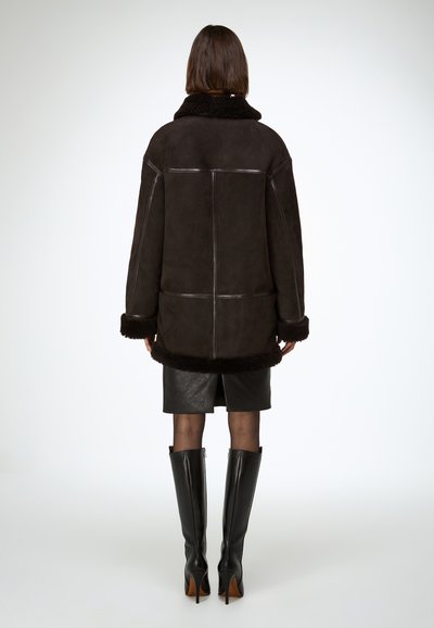 Veste en daim marron avec col et poignets en fausse fourrure, présentant une coupe décontractée et des coutures visibles. Portée avec une jupe en cuir noire et des bottes hautes jusqu'aux genoux.