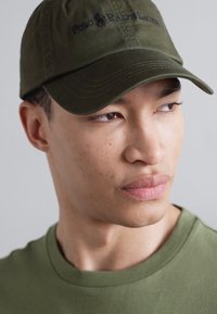 Căciulă de baseball verde măsliniu cu logo-ul brodat "Polo Ralph Lauren", având o vârf curbat și din material de bumbac, purtată cu o cămașă asortată.