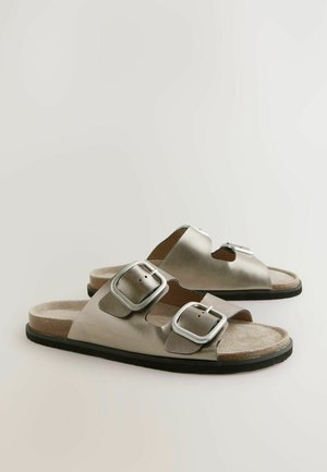 Zilverkleurige metallic slide sandalen met twee verstelbare gespen, een gevormde voetbed en een gestructureerde beige binnenzool, geplaatst op een zwarte rubberen zool.