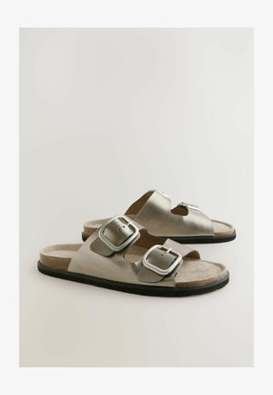 Strieborné metalické sandále na šmyk s dvoma nastaviteľnými prackami, tvarovanou stielkou a textúrovanou béžovou vnútornou stielkou, umiestnené na čiernej gumovej podrážke.