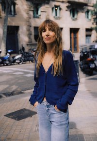 Blå strikket cardigan med V-udskæring og knaplukning, kombineret med lyse denimjeans i et afslappet udendørs miljø.