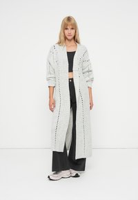 Long gilet gris avec devant ouvert, texture côtelée et détails en œillets. Porté sur un crop top noir et associé à un pantalon noir ample.