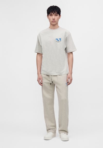 Homme aux cheveux courts noirs, debout, portant un t-shirt à manches courtes gris clair et un pantalon de survêtement assorti avec des baskets blanches sur un fond uni.